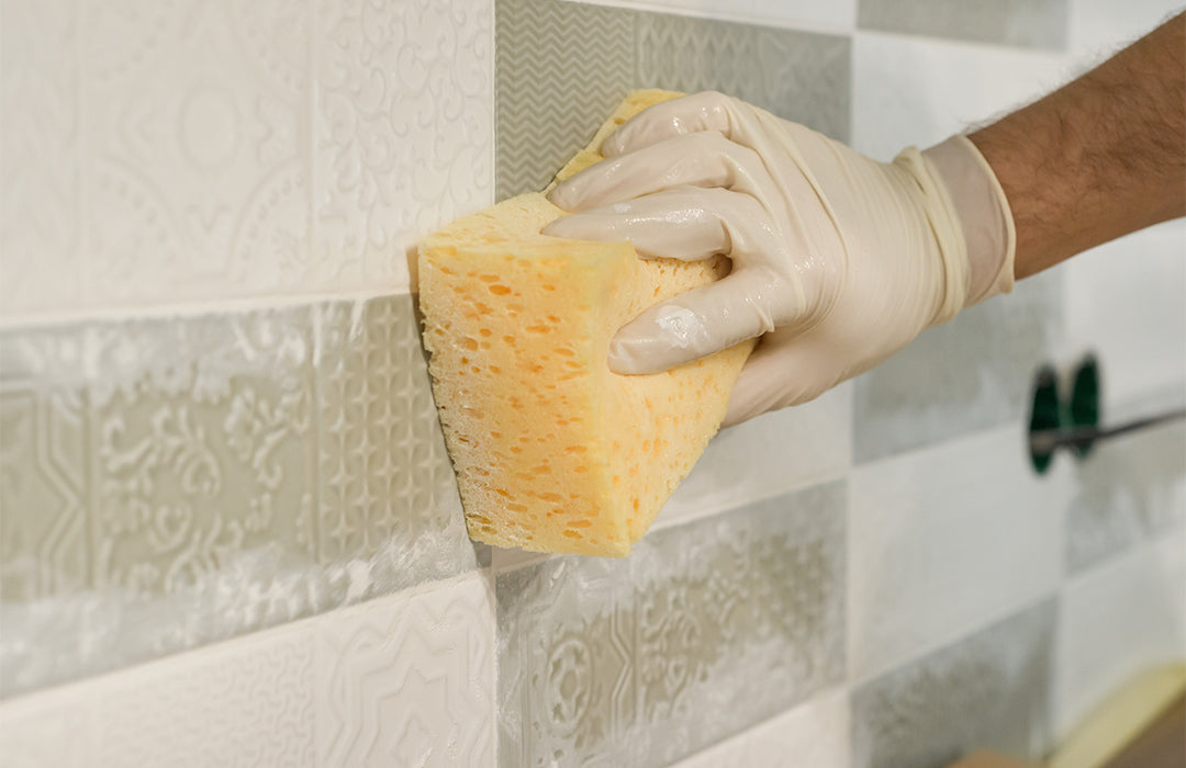 Hand wearing a glove holding a yellow sponge against a tiled wall.