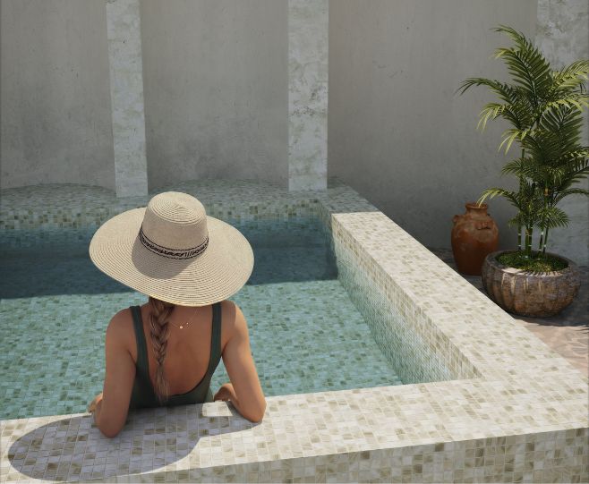 A woman wearing a wide-brimmed straw hat and a dark swimsuit sits at the edge of a tiled swimming pool, viewed from behind. The scene is set in a sunlit outdoor patio area with a potted palm and textured walls.