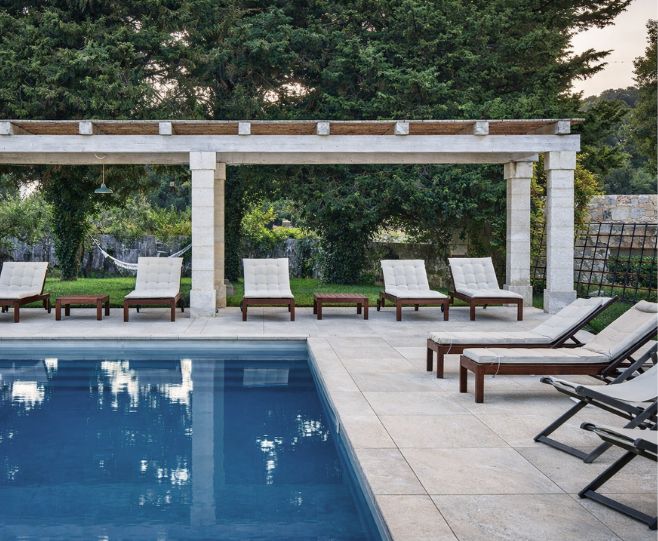 Outdoor poolside patio scene with a white pergola, several lounge chairs, and dense green trees in the background.
