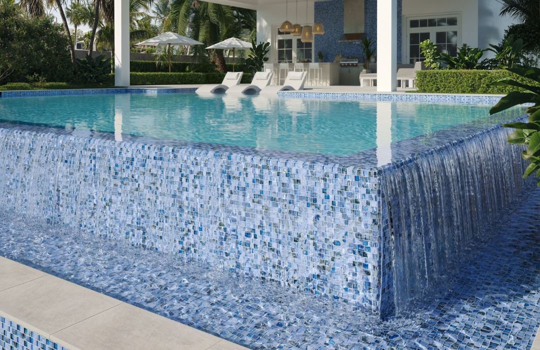 glass pool tile with in pool loungers, outdoor kitchen and umbrellas