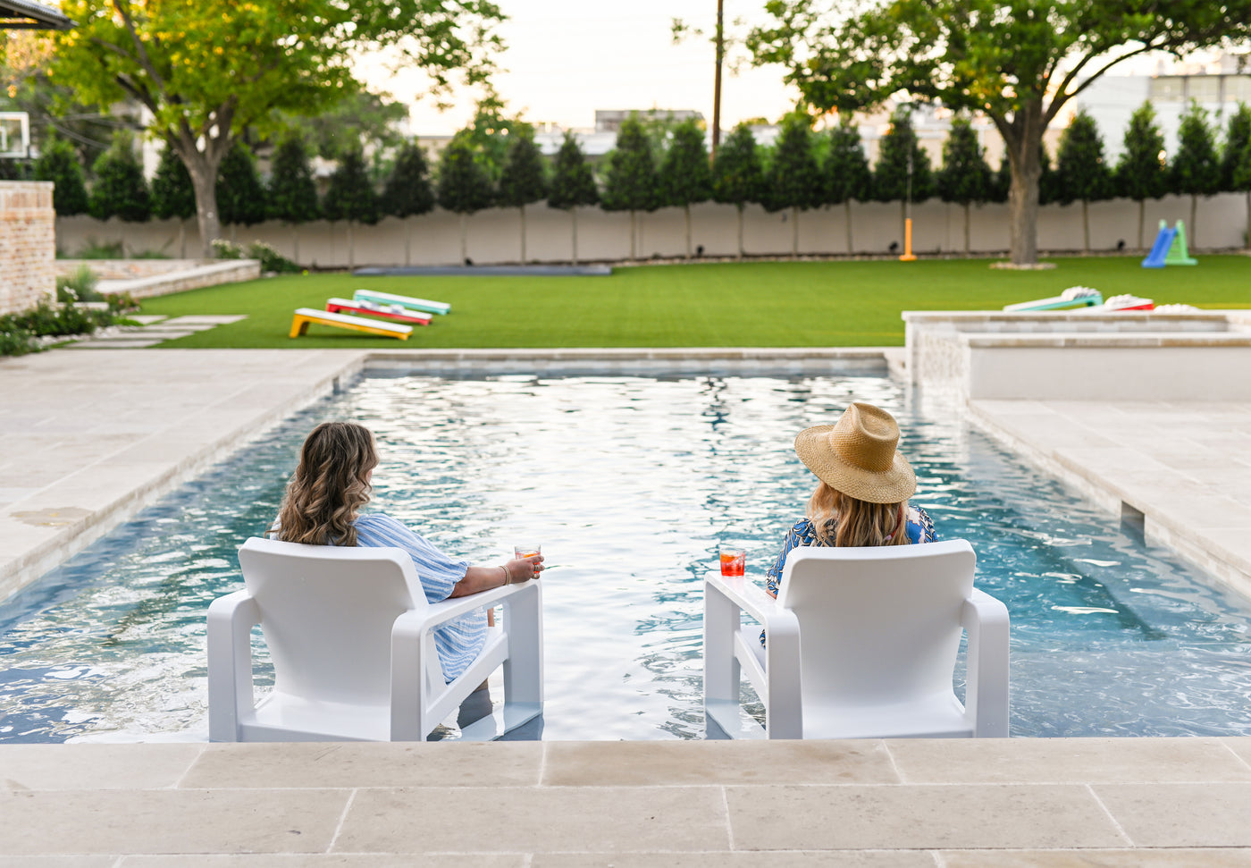 Two people sitting in a pool on Tupelo Goods In-pool lounge chairs in a backyard with green grass and trees.