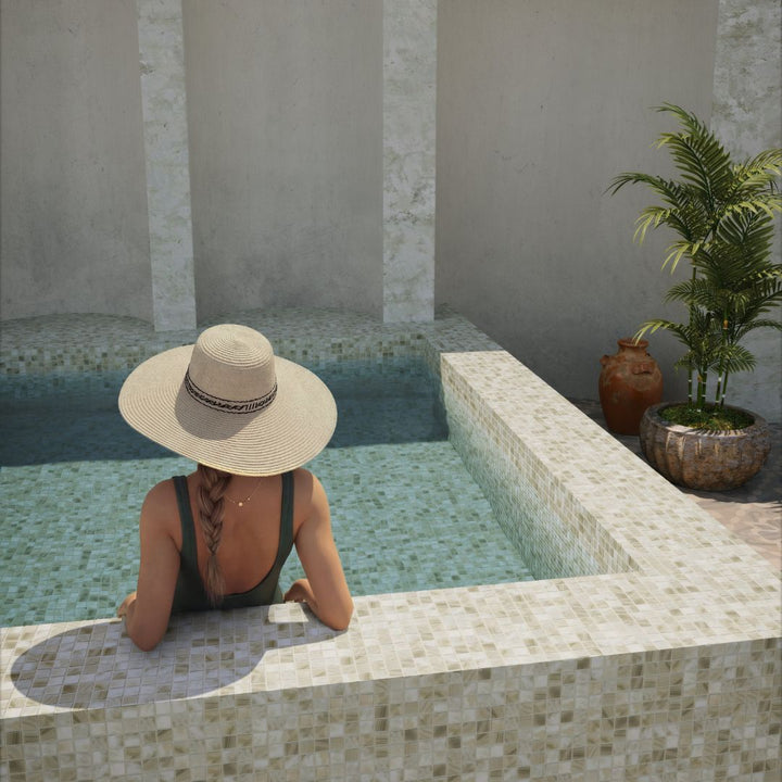 A woman wearing a wide-brimmed straw hat and a dark swimsuit sits at the edge of a tiled swimming pool, viewed from behind. The scene is set in a serene outdoor patio environment with a potted palm and textured walls.
