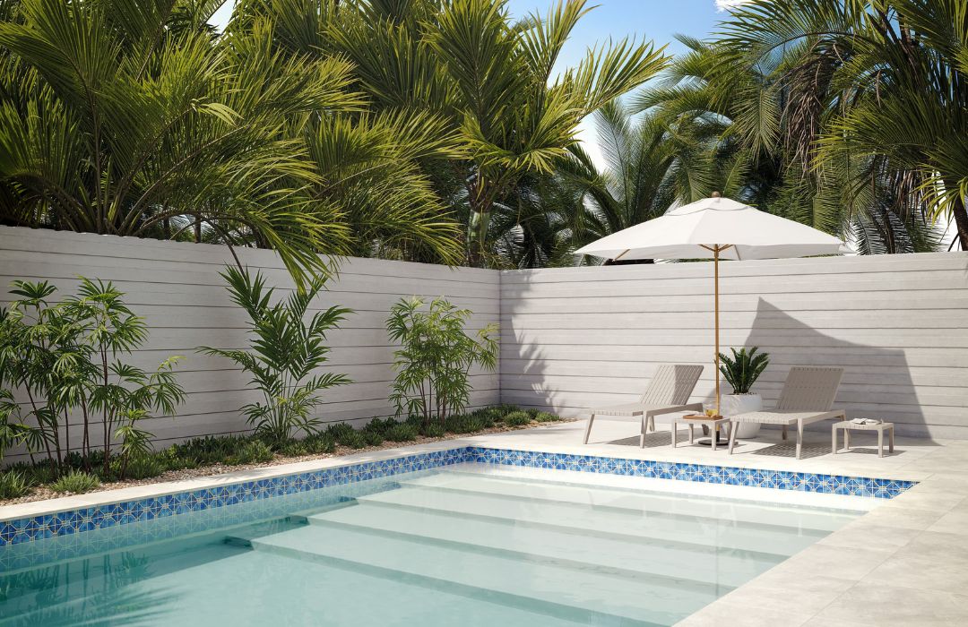 Pool area with lounge chairs, umbrella, and tropical plants against a white fence.
