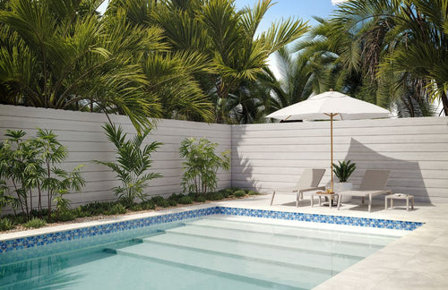 Pool area with lounge chairs, umbrella, and tropical plants against a white fence.